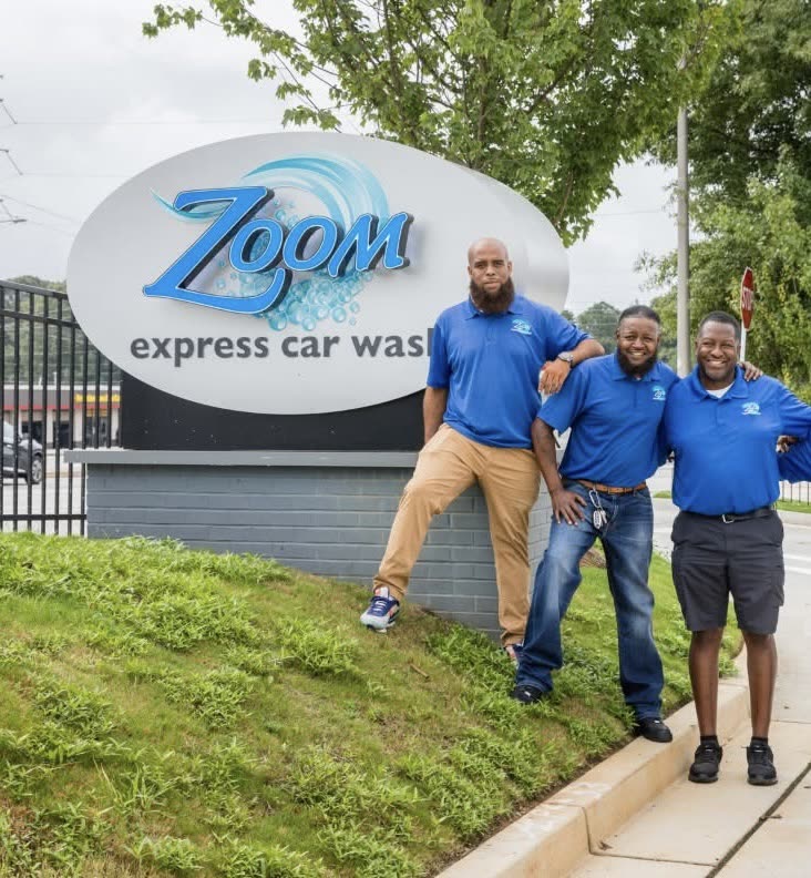 Three Zoom Express team members in blue polos laughing beside the Zoom Express Car Wash sign