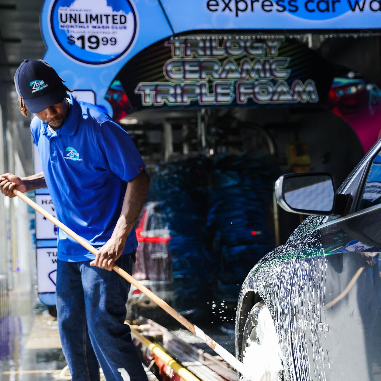 A Zoom Express team member in a branded cap scrubbing a car with a long brush under the Zoom arch